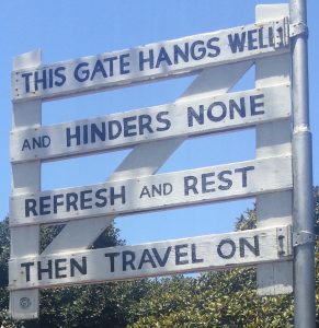 Photo of a white slatted gated with the words painted on it: "THIS GATE HANGS WELL / and HINDERS NONE / REFRESH and REST / THEN TRAVEL ON