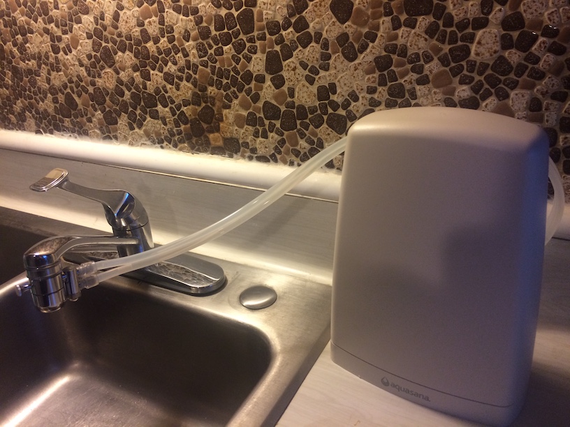 Photo of sink and faucet with with two clear tubes coming from the faucet aerator to a opaque white plastic container on the counter.