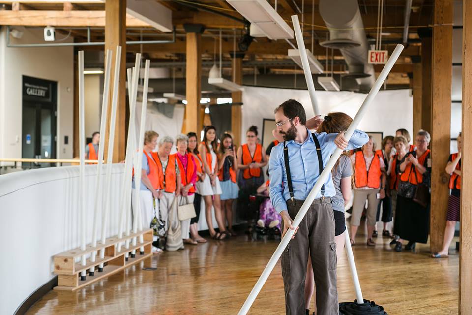Photo of the interior of an art museum. Foreground: Matthew Collie faces camera standing back to back with a woman. Both are holding white PVC poles. Background: Crowd in orange vests watching. Photo by Kensie Wallner.
