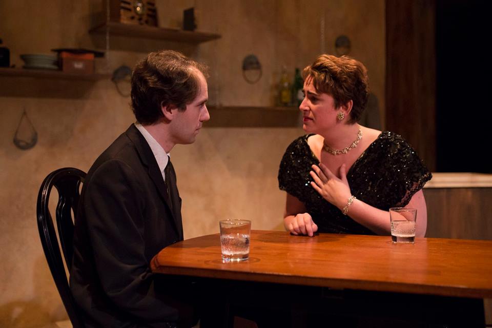 Production photo of The Sweet New by Raymond Rea. Larry, played by Matthew Collie, sits at a table facing Vivian who clutches at her neck worried. Photo by Kensie Wallner.