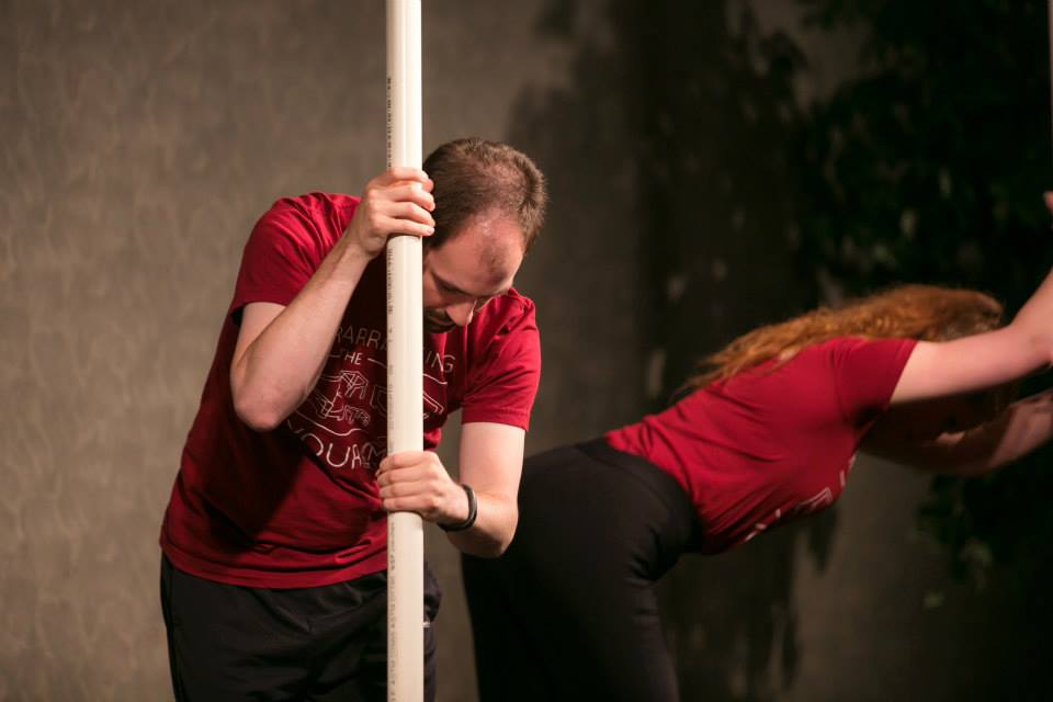 Performance still from The Oil Project of Matthew Collie and woman in red. Matthew Collie faces the camera bent over holding a PVC pole. The woman, bent over, faces away from the camera. Photo by Kensie Wallner.