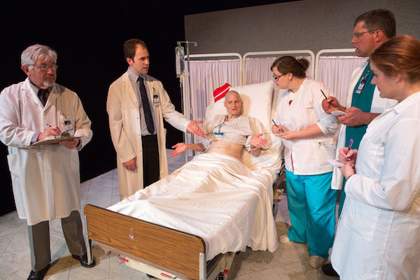 Photo of a hospital bed with a female patient. Standing around the bed are five people dressed in hospital coats or scrubs. To the left is two men, one older, one younger one (Matthew Collie). To the right are two women and one man. Photo by Michael Benedict.