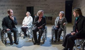 Production photo from 33 Variations. Five people, Matthew Collie, two women, and two men sit in wheelchairs arranged in a row. The men are dressed in 19th century formal attire, the women in modern semi-professional attire. Behind them is a pianist at a piano with scores of music painted on the walls.  Photo by Michael Benedict.